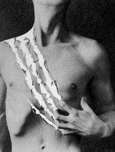 black and white photograph of a shirtless man with torn paper around his chest, looking at the camera