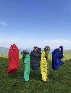 four people in colorful sleeping bags standing on top of a grass covered hill with mountains in the background