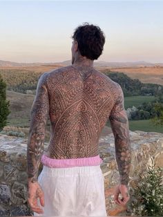 a man with tattoos on his back standing in front of a stone wall and looking at the landscape