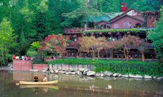 two people in a canoe on the water near a large house with trees and bushes around it