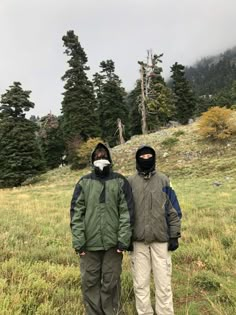 two people standing on top of a grass covered field next to trees and mountains in the background