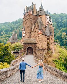 a man and woman holding hands while walking on a stone bridge in front of a castle