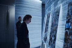 two men standing in front of a wall covered with pictures and papers, looking at each other