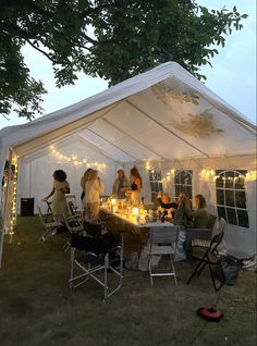 a group of people sitting around a table under a white tent with lights on it