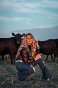 a woman squatting down in front of cows on the grass with her cell phone up to her ear