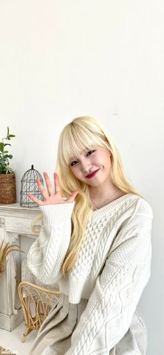 a blonde woman sitting on a chair with her hand up to the camera and wearing a white sweater