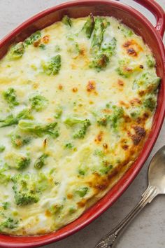 a red casserole dish with broccoli and cheese on top, next to silverware