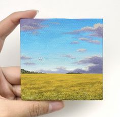 a hand holding up a small square piece of paper with a painting of a field in the background