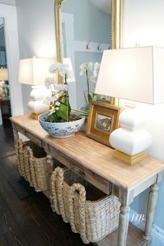 a wooden table topped with a bowl of flowers next to a mirror and lamp on top of a hard wood floor