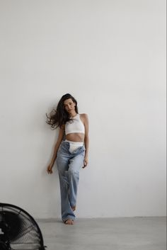 a woman leaning against a wall with her hair blowing in the wind, wearing jeans and a crop top