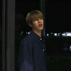 a young man wearing glasses standing in front of a window with the city lights behind him