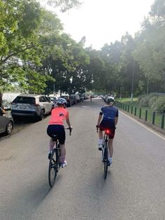 two bicyclists are riding down the street in front of parked cars and trees