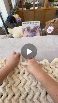 someone is knitting a blanket on the floor in front of a couch with a book about hand knitting