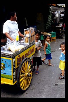 a man selling food from a cart on the street with children standing around him and looking at it