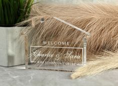 a clear acrylic welcome sign sitting next to a potted plant