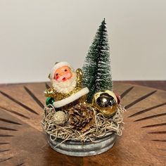 a small christmas decoration with a santa clause sitting on top of a wooden table next to a pine tree