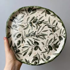 a hand holding a plate with olives painted on the front and sides, against a gray background