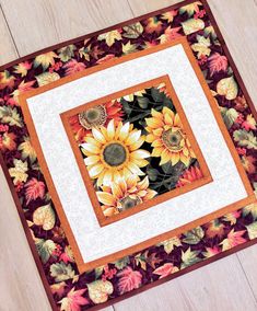 a quilted table topper with sunflowers and leaves on it, sitting on a wooden floor