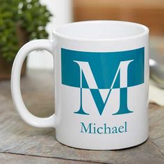 a white coffee mug with the letter m on it sitting on a table next to a potted plant