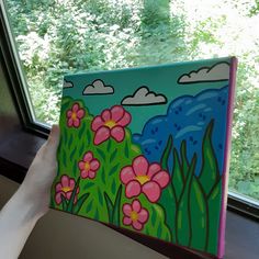 a person holding up a painting in front of a window with trees and bushes outside