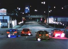 three cars driving down the road at night time with bright lights on them and buildings in the background