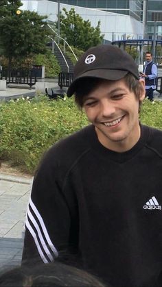 a young man smiles while sitting on a bench in front of a building with people walking around