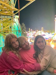 three women sitting on a bench in front of a carnival ride at night with ferris wheel in the background