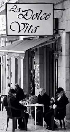 three men sitting at a table in front of a building with a sign that says la dolce vita