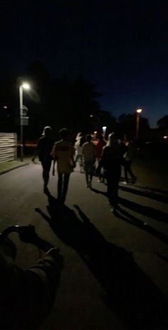 a group of people walking down a street next to a light pole at night with their shadows on the ground