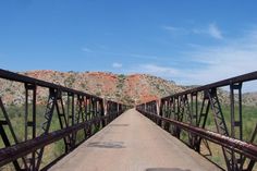 an old rusty bridge crosses over a dirt road in front of mountains and grass on a sunny day