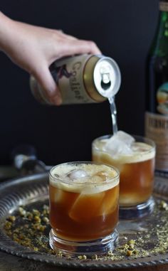 two glasses filled with iced tea being poured from a can into the glass on top of a metal tray