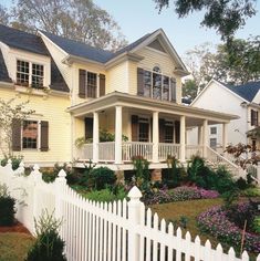 a white picket fence in front of a yellow house with lots of flowers on the lawn