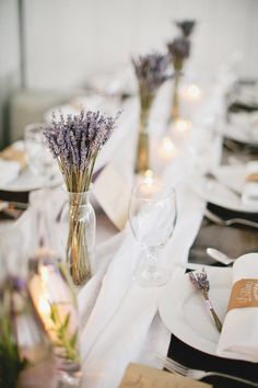 the table is set with white plates, silverware and lavenders in vases