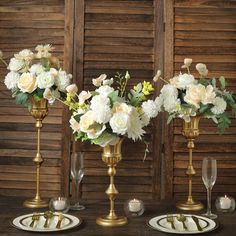 three gold vases filled with white and yellow flowers on top of a wooden table