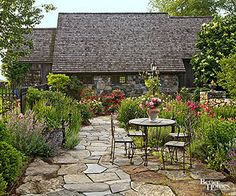 a stone path leads to an outdoor dining area in front of a house with flowers and greenery