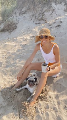 a woman sitting in the sand with her dog and holding a coffee cup next to her