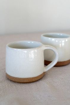 two white coffee mugs sitting on top of a beige cloth covered table next to each other