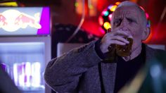 an older man drinking from a beer glass in front of neon signs at night time