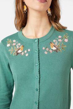 a woman wearing a green cardigan with birds and flowers on the front, standing in front of a white background