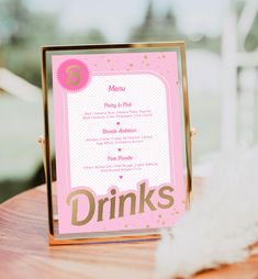 a pink and gold drink menu sitting on top of a wooden table next to a white feather