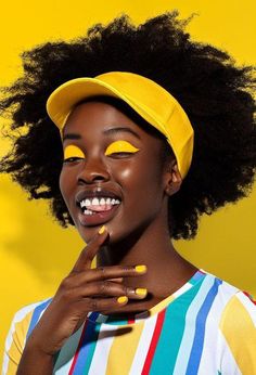 a woman with yellow painted on her face wearing a baseball cap and striped shirt, posing in front of a yellow background