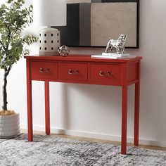 a red table with two drawers in front of a painting and potted plant next to it