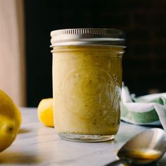 a jar filled with yellow liquid next to two lemons and a spoon on a table