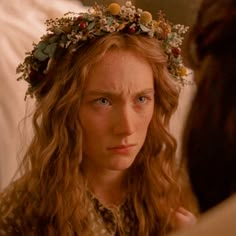 a woman with long hair wearing a wreath of flowers on her head looking at another woman