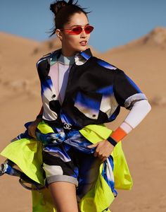 a woman wearing sunglasses and a colorful dress in the middle of sand dunes with her legs spread out