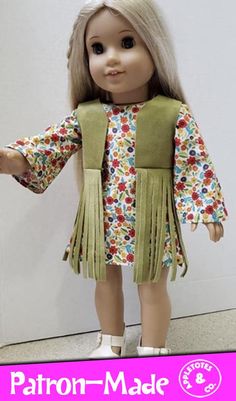 a doll with blonde hair wearing a green dress and fringed jacket, standing in front of a white wall