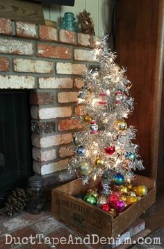 a small christmas tree in a box next to a fire place with ornaments on it