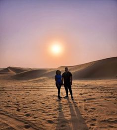 two people standing in the middle of a desert with the sun setting over sand dunes