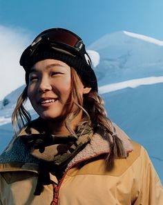 a woman wearing ski goggles standing in front of a snow covered mountain with her eyes closed