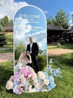 a man and woman standing in front of a welcome sign with flowers on the grass